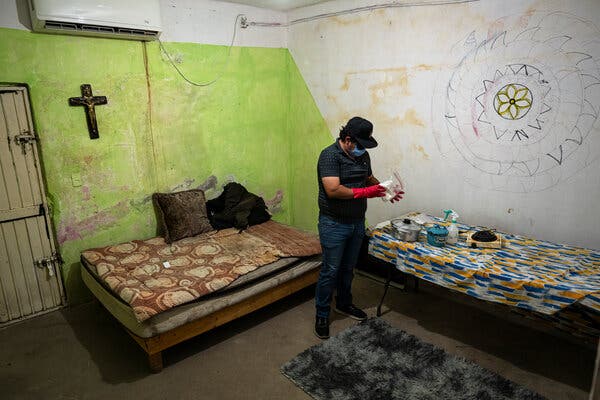 A man wears red gloves while mixing chemicals in a sparse room with a cross on the wall. 