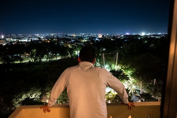 A man in a white shirt looks out over a patio at a city at night.