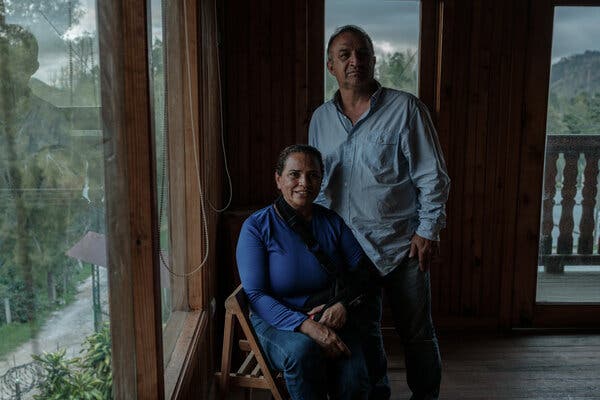 Un hombre con camisa azul de pie detrás de su esposa, que está sentada.