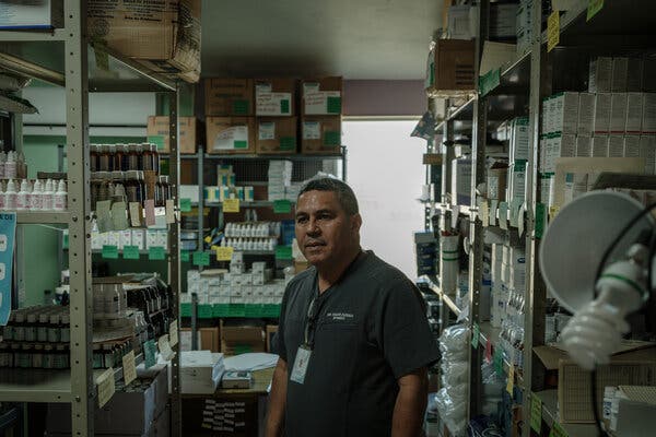 Un hombre de camiseta oscura parado en medio de estantes con medicamentos.