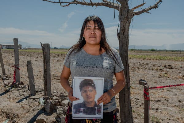 Una mujer, con una camiseta gris, sostiene una foto de un hombre joven. Está de pie delante de un campo árido.