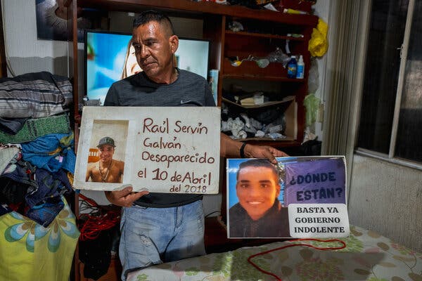 Un hombre, de pie ante unas estanterías llenas de enseres y un televisor, sostiene dos carteles caseros, cada uno con la foto de un joven.