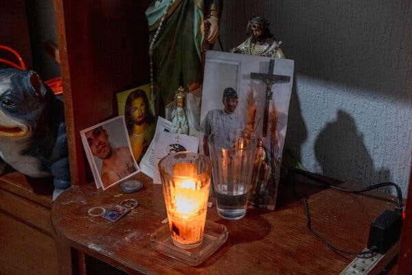 Una vela votiva, imágenes y estatuillas de Jesús y fotografías de un joven, sobre un estante de madera.