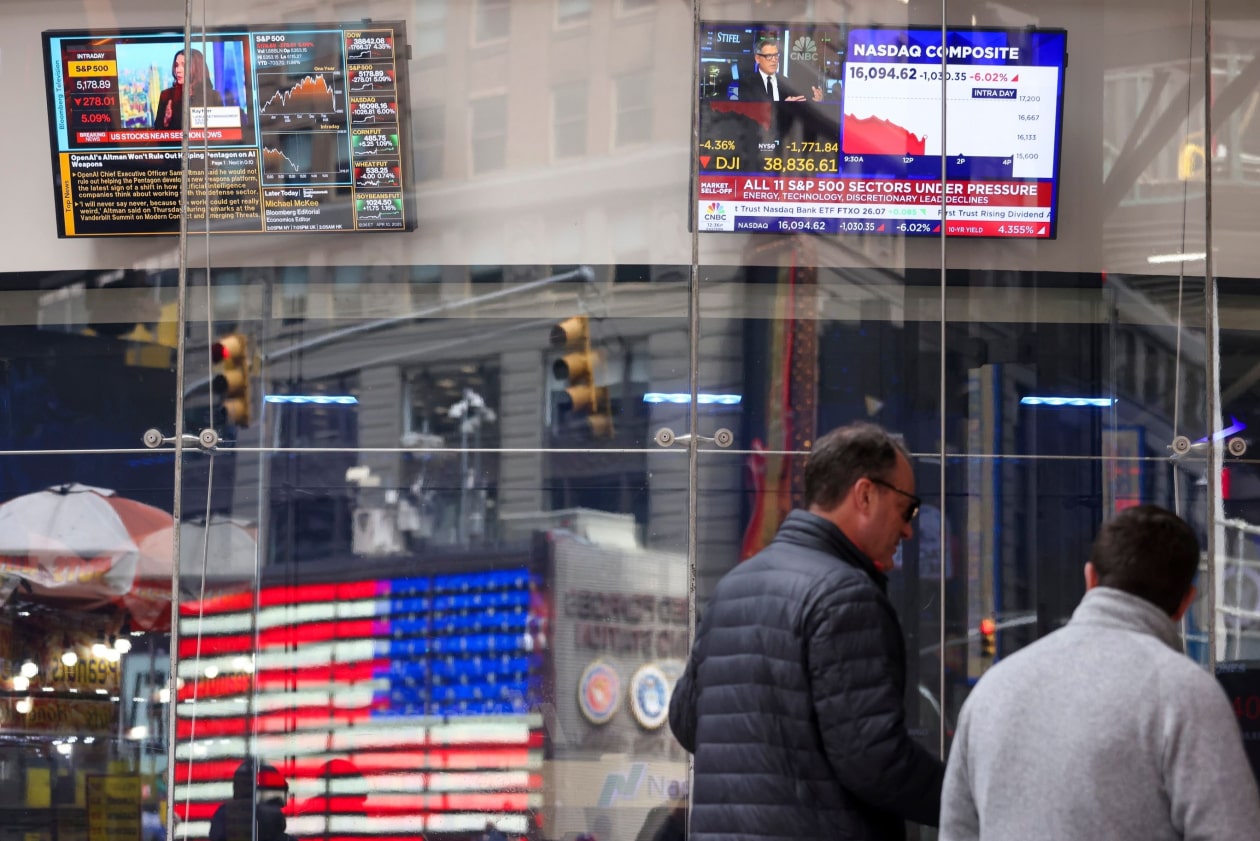 La gente pasa frente a una oficina del Nasdaq en Times Square; las pantallas muestran informes de noticias sobre acciones con tendencia a la baja.