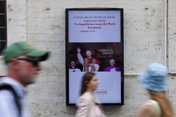 La gente pasa junto a una pantalla digital en la que se ve la imagen del papa saludando con la mano.