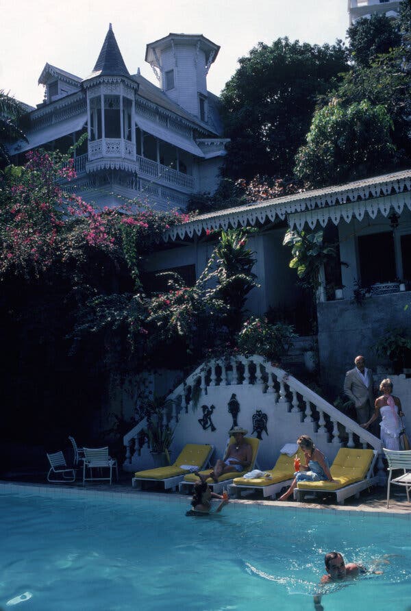 Gente que nadaba en una piscina, mientras otros se sentaban en tumbonas. Al fondo se ve parte de una estructura del edificio con ventanas amplias y celosías en los balcones.