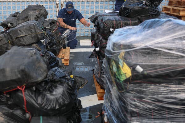 Un miembro de la Guardia Costera arrodillado en el suelo de un barco en medio de grandes pilas de carga en bolsas de basura negras.