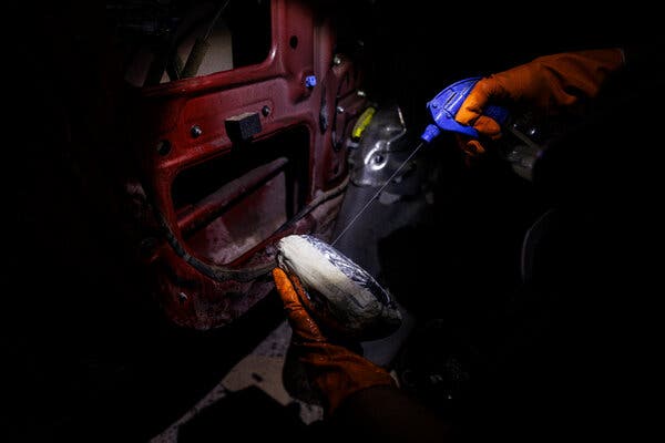 Una persona con guantes anaranjados sostiene una botella con un rociador azul mientras apunta hacia un paquete.