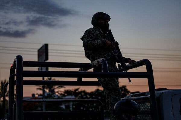 Un hombre con equipo militar y un arma larga vigila al anochecer.