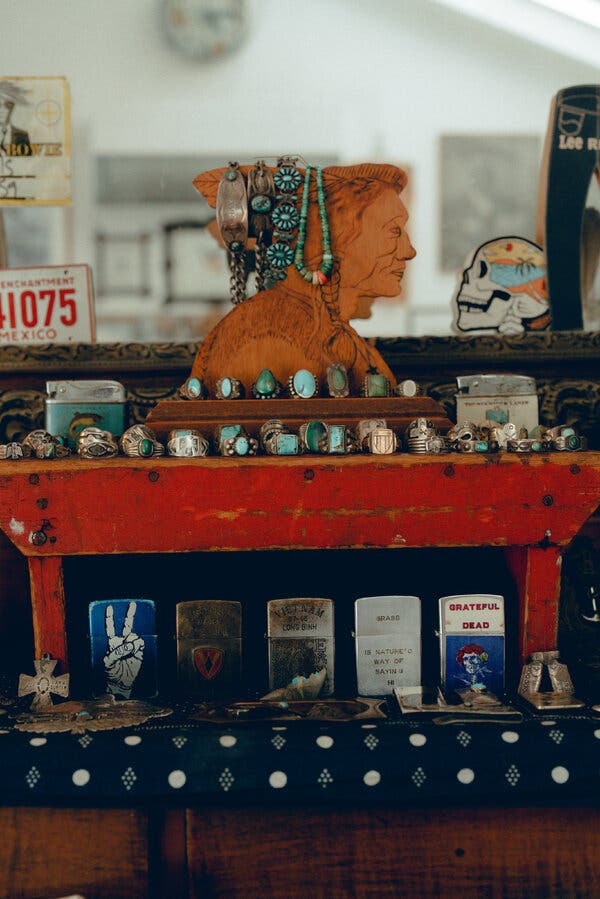 Una cómoda de madera roja con un busto de una figura nativa está llena de anillos y joyería de color turquesa, y encendedores de metal en la parte inferior.
