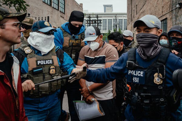 Personas migrantes esposadas se abrían paso entre una multitud de manifestantes en Chicago. Funcionarios de migración con máscaras y chalecos con parches en los que se lee “Policía HSI” se situaban a ambos lados y detrás de los migrantes.