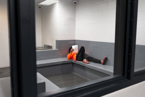 Un migrante hondureño vestido con camisa naranja, pantalones negros y sandalias marrones yace en una celda de detención desierta.