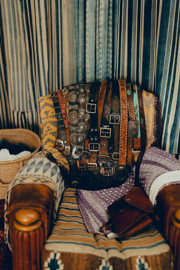 Un sillón de cuero marrón cubierto de mantas con motivos geométricos. Sobre el respaldo hay muchos cinturones.
