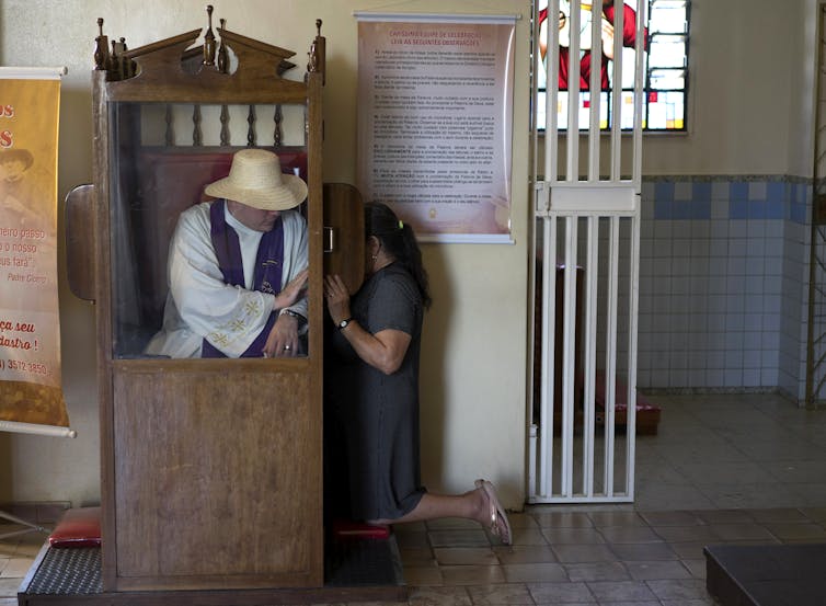 Un hombre con una túnica blanca y un sombrero de paja está sentado dentro de un cubículo de madera mientras una mujer se inclina junto a él, con una pequeña pantalla protegiendo su rostro.