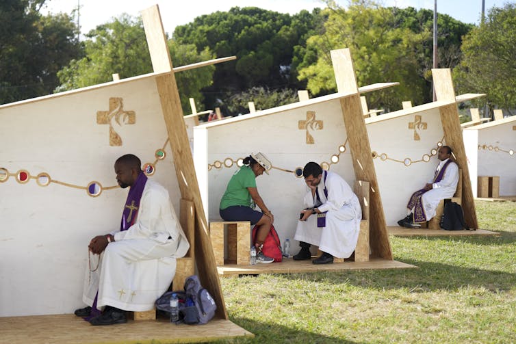 Una hilera de estructuras al aire libre, con tres paredes para mayor privacidad, dispuestas en fila, con un hombre vestido de blanco sentado en cada una.