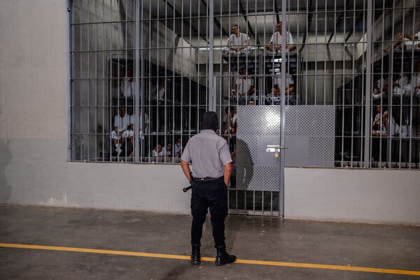 Un guardia de prisiones vigilaba a unos hombres con uniforme blanco sentados en literas en una celda grande.