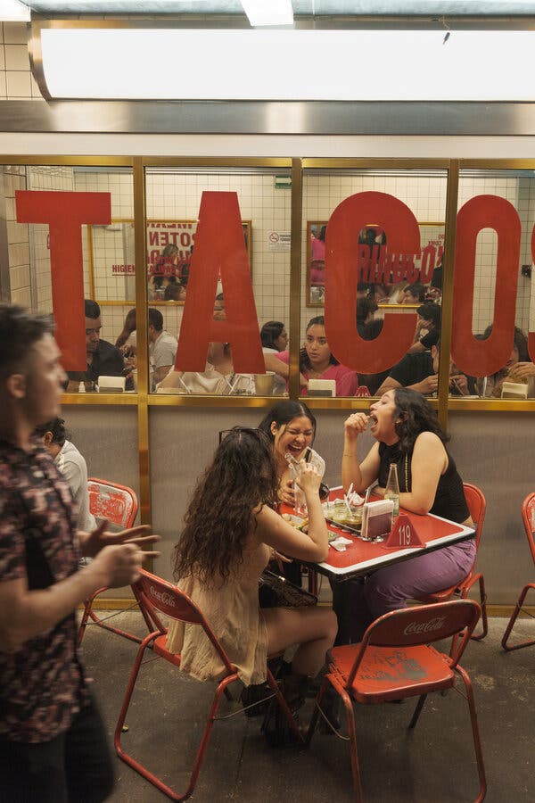 Una zona de asientos al aire libre con gente sentada en pequeñas mesas y sillas rojas frente a un restaurante con la leyenda “TACOS” escrita en grandes letras rojas en la ventana. 