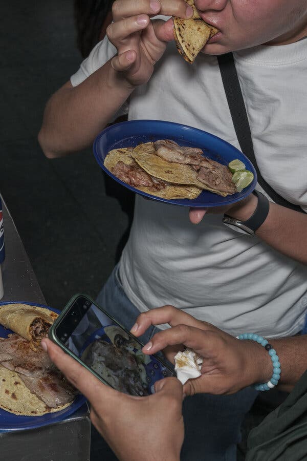 Una persona sostiene un plato azul con tacos de carne y limones a un lado, mientras otra le toma una foto con un teléfono a su plato.