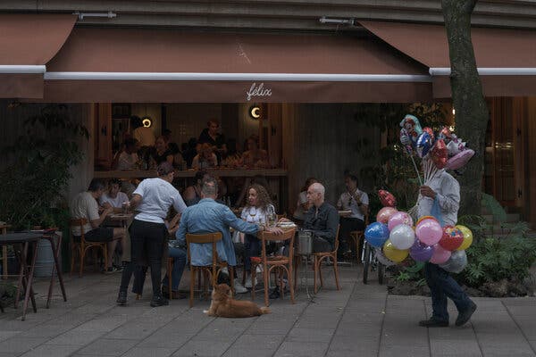 La gente está cenando en mesas al aire libre bajo un toldo marrón que dice “félix”, mientras que una persona que vende globos de colores y un perro están en la acera en primer plano.