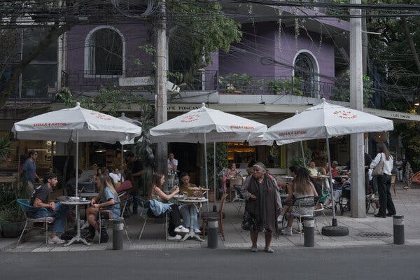Una mujer pasa junto a varias mesas de cafés al aire libre con gente sentada bajo grandes sombrillas blancas frente a un edificio morado.