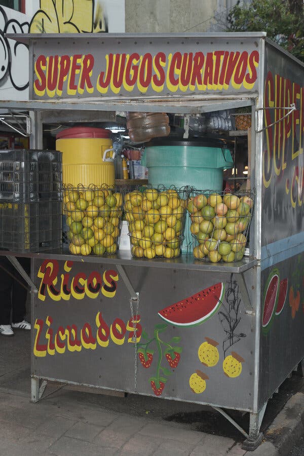 Un puesto callejero de comida con la leyenda “SUPER JUGOS CURATIVOS” pintada en la parte superior, y tres cestas de naranjas sobre su barra lateral.

