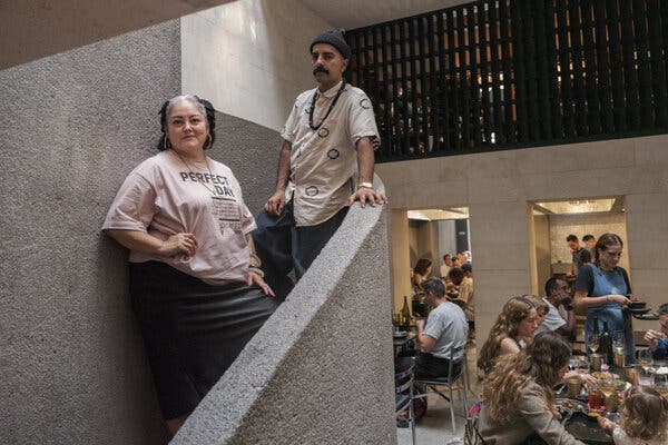 Una mujer con una camiseta rosa y un hombre con bigote y gorro están de pie en una escalera de hormigón con vistas a un concurrido comedor en la parte de abajo. 