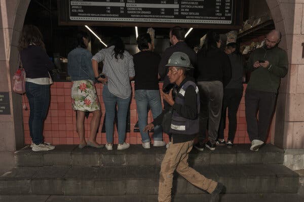 Un hombre con casco y chaleco camina en primer plano frente a una fila de personas que esperan para pedir en un mostrador. 