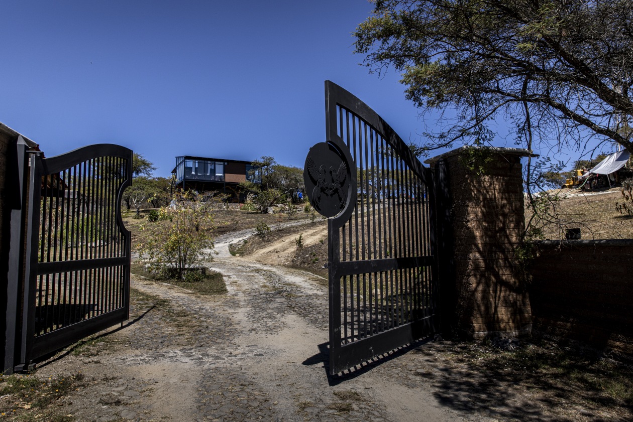 Una puerta metálica abierta con un emblema y un camino de tierra que conduce a una cabaña moderna.
